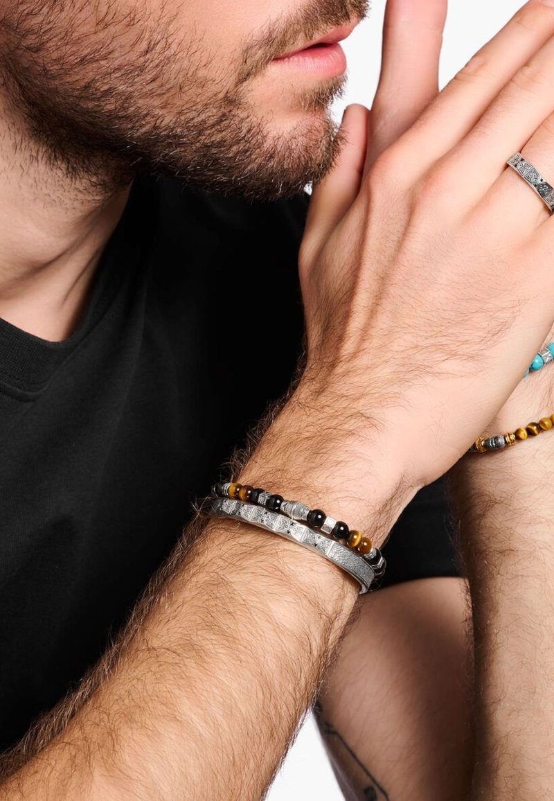 Primo piano di un uomo con barba che indossa una camicia nera, con le mani vicino al viso, adornato con un anello d'argento lavorato e due braccialetti di perline.