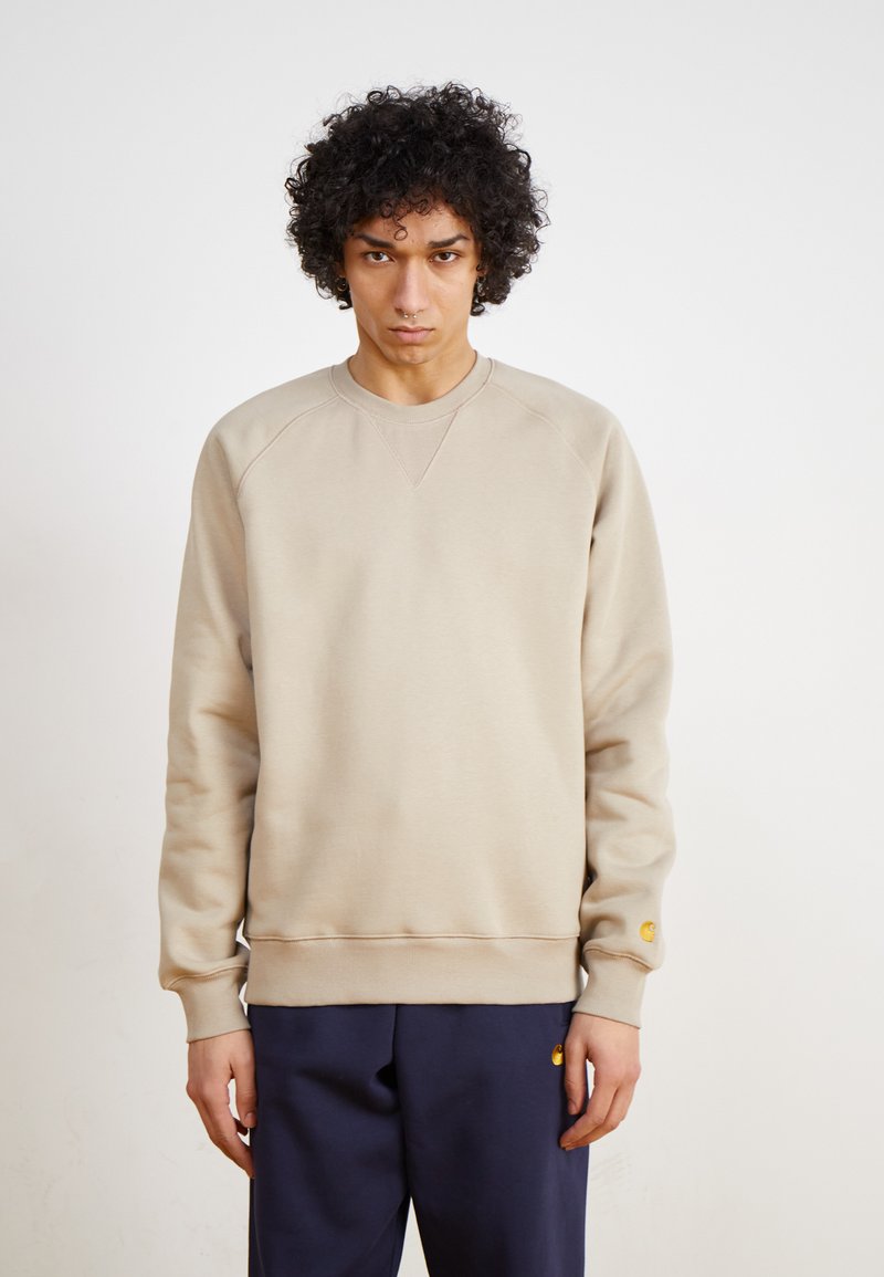 Beige sweatshirt med raglanærmer, ribbede manchetter og bund. Har et lille gult logo på venstre ærme. Bæres med marineblå bukser.