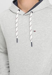 Felpa grigia realizzata in un tessuto morbido, con cappuccio regolabile dotato di dettagli a righe e un piccolo logo sul petto.