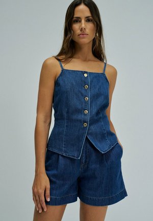 Femme portant un haut en denim sans manches à boutons et un short en denim assorti, debout devant un fond uni clair.