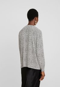 Schwarz-weiße gepunktete Bluse mit langen Ärmeln, lockerer Passform, Rundhalsausschnitt und Faltendetail im Rücken. Glatter Stofftextur.