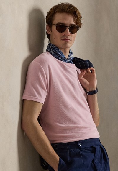 Άνδρας που φοράει στρογγυλά γυαλιά ηλίου, ροζ μπλούζα με κοντά μανίκια, μπλε κασκόλ με σχέδια, σκούρο μπλε παντελόνι και ρολόι, ακουμπώντας στον μπεζ τοίχο.