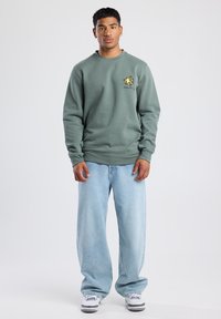 Green sweatshirt, soft fabric, crew neck, ribbed cuffs, logo detail on chest. Paired with light blue baggy jeans and white sneakers.