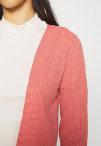 Červený žebrovaný cardigan s otevřeným předním dílem, s dlouhými rukávy. Nosený přes světlý rolák, který předvádí jemnou texturu.