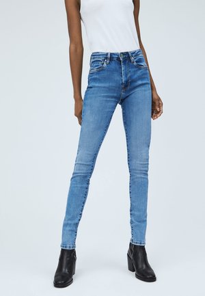 Nainen, joka on pukeutunut vaaleansinisiin kapeisiin farkkuihin, valkoiseen hihattomaan paitaaan ja mustiin korkoankkureihin, seisoo vaaleaa taustaa vasten.