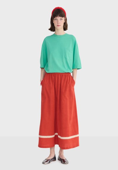 Jeune femme debout portant une chemise verte oversize, un pantalon large rouge avec bordure blanche, un bandeau rouge et des chaussures plates argentées sur un fond uni.