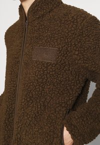 Personne portant une veste polaire marron à zip avec une surface texturée et un écusson sur la poitrine gauche. Main dans la poche latérale.