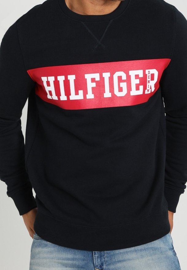 Felpa blu navy con banda rettangolare rossa e logo "HILFIGER" in bianco. Presenta maniche lunghe e polsini a costine. Tessuto morbido al tatto.
