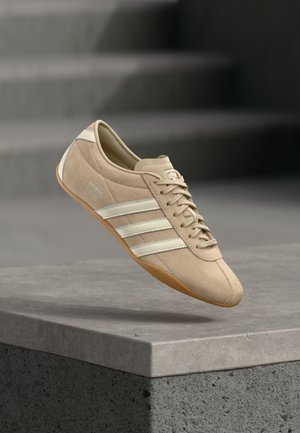 Light beige sneaker with three white horizontal stripes, suede upper, and a rubber gum sole, featuring a logo on the side.