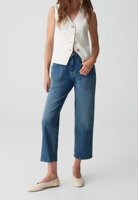 Vit linne ärmlös väst med knappar, tillsammans med högmidjade blå denimjeans. Modellen bär krämfärgade ballerina-skor, vilket visar en avslappnad outfit.