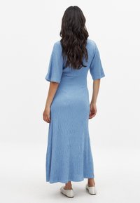 Blauwe, gestructureerde maxi-jurk met korte, uitlopende mouwen en een getailleerde bodice, die uitloopt in een iets uitlopende zoom. Gdrag met witte instappers.