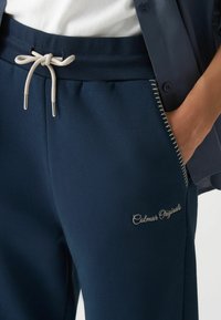 Tamnoplave trenirke od mekanog materijala, imaju elastičnu waistband s bež vezicom i izvezenim logotipom "Colmar Originals".