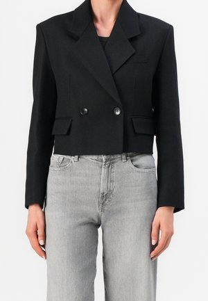 Frau trägt einen schwarzen, taillierten, doppelt geknöpften Blazer mit breiten Revers und graue, hoch taillierte Jeans vor weißem Hintergrund.