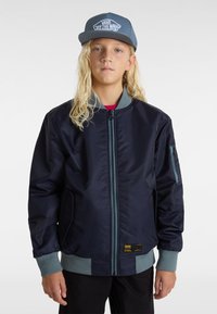 Chaqueta de piloto náutica con cremallera frontal, puños acanalados y bolsillos laterales. Presenta detalles en gris y un bolsillo con cremallera en la manga. Lleva puesta una gorra.
