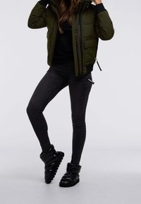 Veste en olive vert bouffant avec des accents noirs, portée avec un jean skinny noir foncé et des bottes noires brillantes, montantes jusqu'à la cheville, avec une semelle striée.