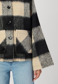 Veste en laine mélangée avec un motif à carreaux noir et beige, dotée d'une fermeture à bouton, de deux poches avant et de manches larges. Finition texturée.