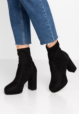 Sorte ruskind ankelstøvler med blokhæle, båret med cropped blå jeans mod en ensfarvet hvid baggrund.