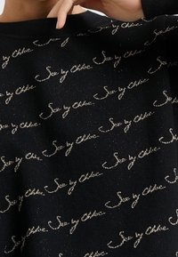 Zbliżenie na czarną dzianinę z powtarzającym się odręcznym napisem "Sea by Chloé" wykonanym błyszczącą nicią, noszoną przez osobę z ręką blisko szyi.