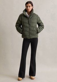 Veste rembourrée verte avec col haut et fermeture éclair à l'avant ; portée sur un pantalon noir ajusté et des chaussures beiges, présentant une finition texturée et un design matelassé.