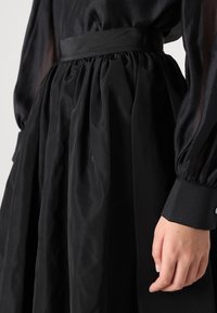 Juoda suknelė su plišine sijonu, elastic juosmeniu ir ilgomis permatomomis rankovėmis. Audinys su tekstūra ir švelniu blizgesiu. Detalė su užsegamais rankogaliais.
