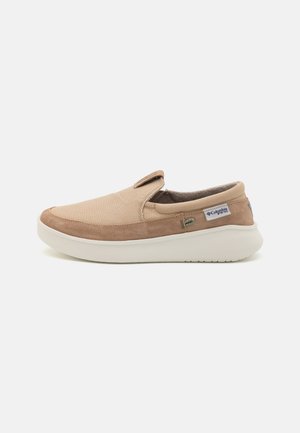 Beige slip-on sko med kanvasoverdel, semskede detaljer, polstret såle og en diskret merkevarelogo på siden.