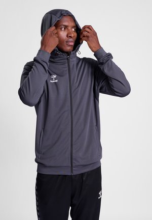 Hooded sportjack in donkergrijs, met een rits aan de voorkant, gestructureerde stof, logo op de borst, met verstelbare capuchon en zakken. Gecombineerd met zwarte broek.