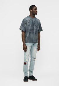 Homme debout portant une chemise grise délavée et surdimensionnée avec le chiffre "99" et des lettres, un jean clair déchiré avec des patchs, et des chaussures noires sur un fond blanc.