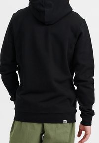 Schwarzer Kapuzenpullover mit weicher Textur, ausgestattet mit einer Kängurutasche und einem kleinen Logopatch am Saum, kombiniert mit hellgrünen Hosen.