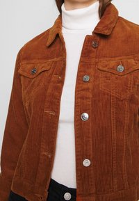 Bruine corduroy jas met zilveren knopen, gedragen over een witte geribde turtleneck en zwarte broek.