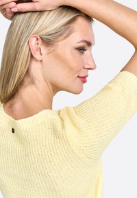 Maglione giallo lavorato a maglia con maniche corte, caratterizzato da un motivo testurizzato e un piccolo accento nero vicino al colletto.