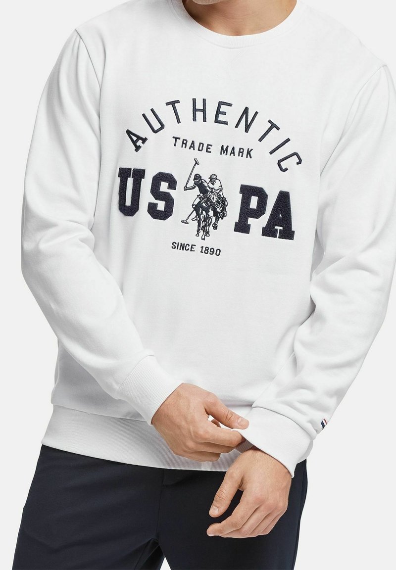 Homme portant un sweat-shirt blanc avec l'inscription "AUTHENTIC US PA" et un graphisme de joueur de polo sur le devant, ajustant le poignet, sur un fond uni.
