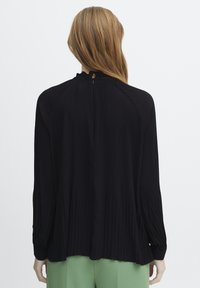 Blouse noire plissée avec des manches longues et un col haut. Présente une fermeture en forme de clé à l'arrière et une texture lisse.
