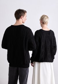 Black knit sweaters with textured cable patterns, loose fit, and ribbed cuffs, worn by two individuals from behind. One wears light-colored pants.