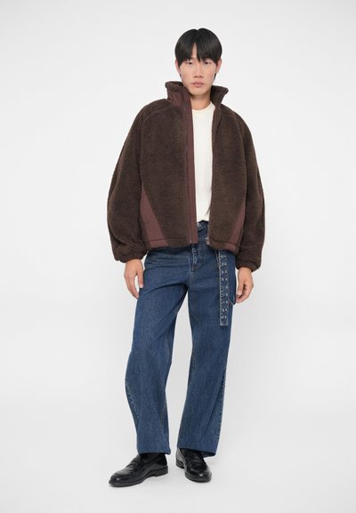 Veste en polaire marron avec un col haut et des panneaux en tissu contrastant, associée à un jean en denim bleu à jambe large et des chaussures noires.
