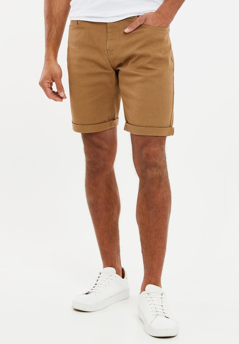 Pantalones cortos de algodón marrón con corte recto, puños doblados y bolsillos laterales, combinados con zapatillas blancas.