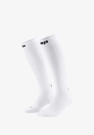 Knee-high white compression socks with ribbed texture, black branding at the top, and patterned grip on the bottom of the foot.