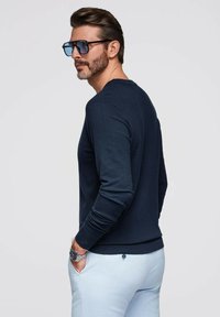 Marineblauer Pullover mit langen Ärmeln, gerippten Bündchen und Saum, kombiniert mit hellblauen Hosen. Zubehör umfasst dunkle Sonnenbrillen und eine silberne Uhr.