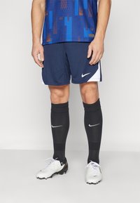 Mørkeblå Nike shorts med hvide accenter, parret med sorte knæstrømper og hvide fodboldstøvler. Har et åndbart materiale og en atletisk pasform.