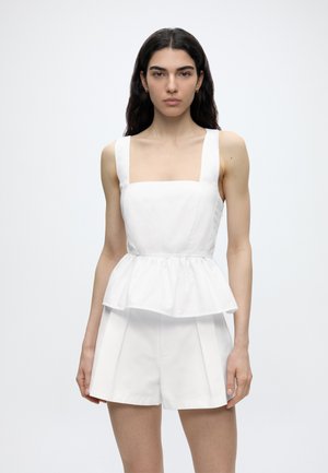 Γυναίκα με μακριά σκούρα μαλλιά, που φοράει λευκό αμάνικο τοπ peplum και ψηλόμεσα λευκά σορτς, όρθια μπροστά σε απλό φωτεινό φόντο.