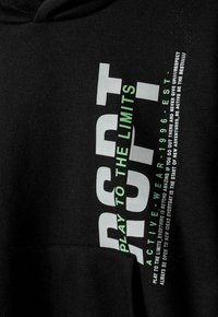 Sudadera negra con bolsillo frontal, con un texto en negrita de color blanco y verde: "RSPT JUEGA HASTA LOS LÍMITES" junto con texto adicional en una fuente más pequeña.
