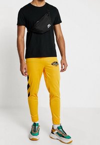 T-shirt noir, pantalon de survêtement jaune avec des rayures noires, et baskets multicolores. Accessoires : un sac bandoulière noir avec logo.
