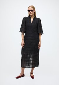 Juoda midi suknelė iš tekstūruotos medžiagos, su V formos iškirpte ir pūstomis rankovėmis. Dėvima su raudonais butais, modelis taip pat nešioja stačiakampius akinius nuo saulės.