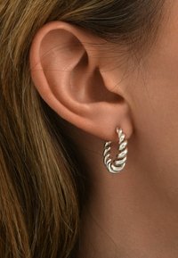 Boucle d'oreille en argent avec un design torsadé, dotée d'une finition polie. Portée sur le lobe de l'oreille, mettant en valeur sa forme ronde et sa texture lisse.