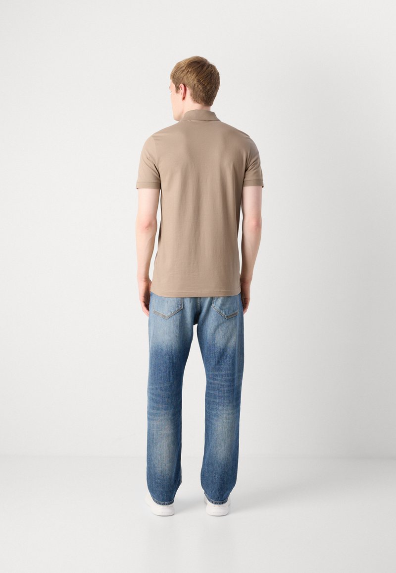 Un jeune homme debout, de dos, portant un polo beige à manches courtes, un jean bleu délavé et des baskets blanches sur fond uni.