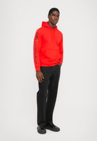 Sudadera roja con capucha y un bolsillo frontal, con una branding negra de EA7 en la manga; combinada con jeans negros de pierna recta y zapatillas.