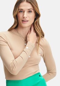 Langsleeve, getailleerd top in beige, gemaakt van soepele stof. Model draagt gelaagde zilveren kettingen. Groene rok zichtbaar. Neutrale achtergrond.