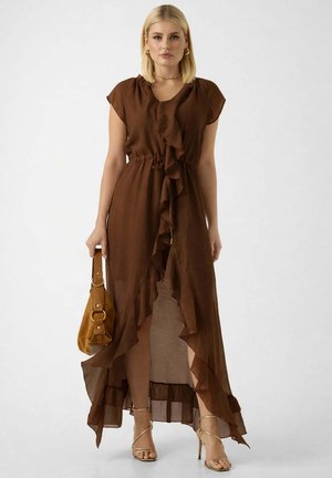 Kvinde med blondt hår iført en brun flæset ærmeløs kjole, der holder en brun håndtaske og gyldne højhælede sandaler med stropper.