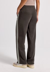 Person som bär mörkgrå hoodiebukser med vita sidostripes och beige sneakers, står med en hand i fickan, vy från bakom.