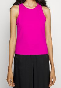 Vrouw met een mouwloze felroze top en losse zwarte broek, zichtbaar van schouders tot heupen tegen een effen achtergrond.