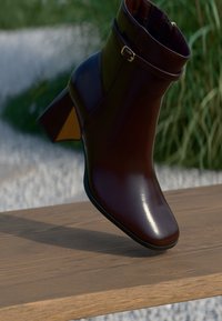 Dunkelbraune Leder-Ankle-Boots mit Blockabsatz und Schnallenriemen, die auf einer Holzoberfläche im Freien ruhen, mit verschwommenem grünem Hintergrund.